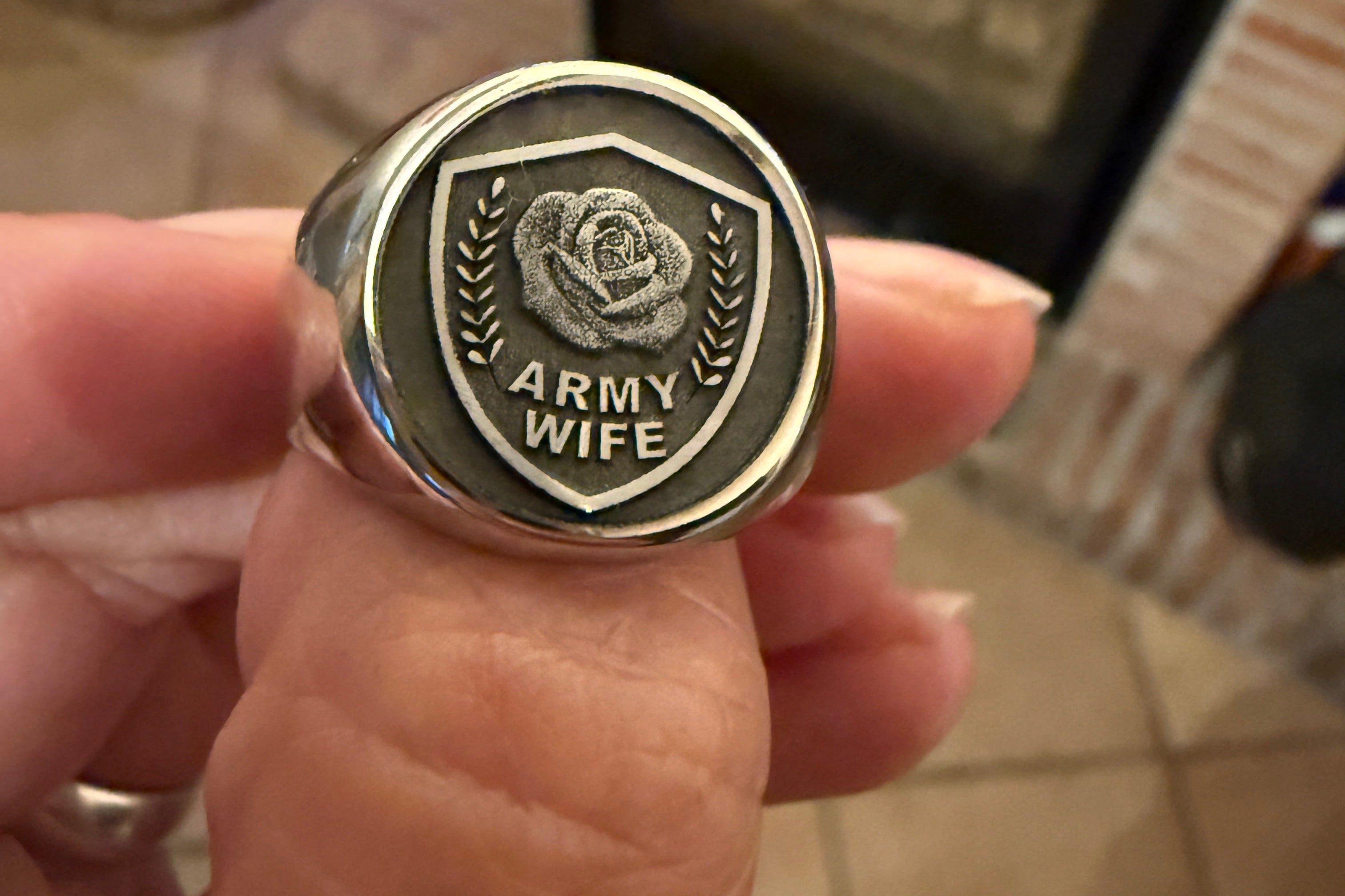 Hand holding a stainless steel signet ring with 'Army Wife' inscription against a home interior background